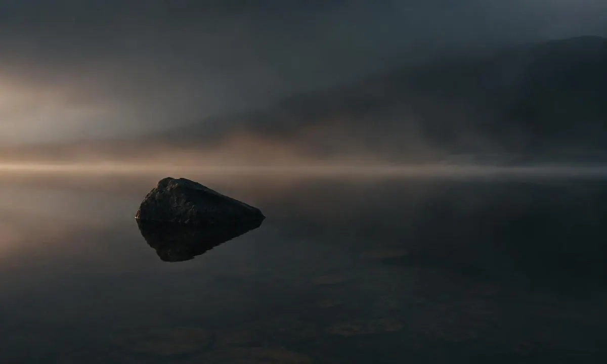 Ein einzelner dunkler Stein ragt aus spiegelglattem Wasser im Nebel